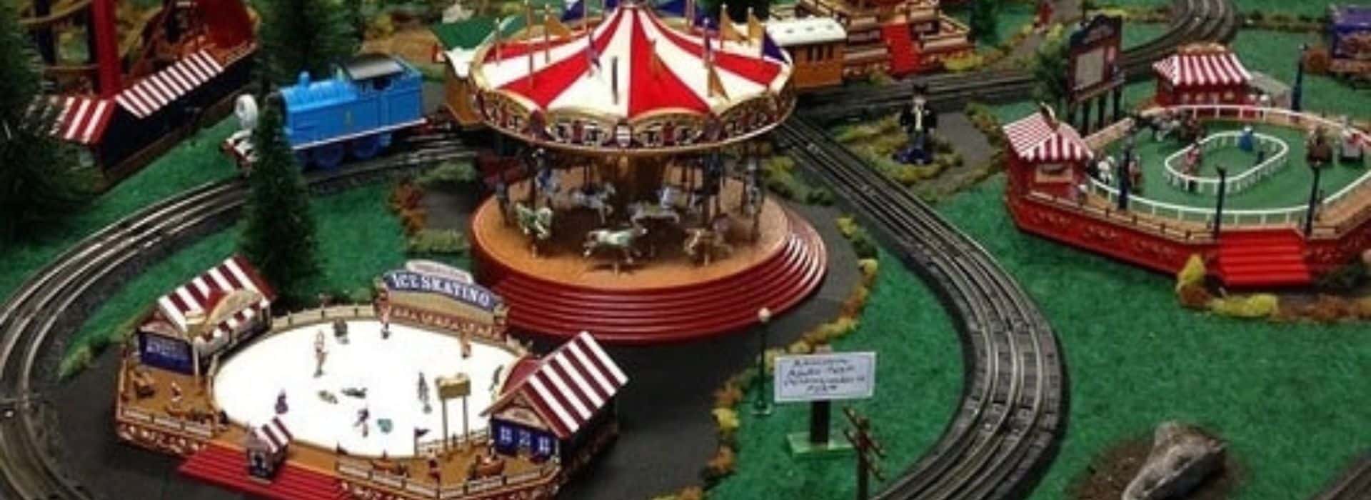 O scale train going by a carousel, ice rink, and horse track.