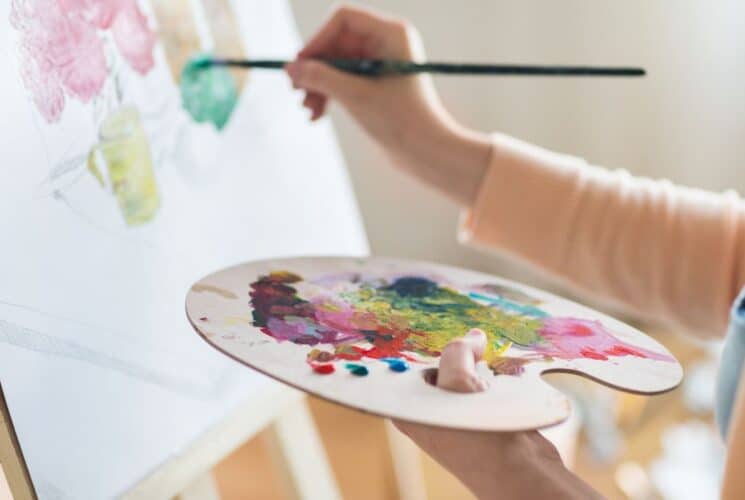 A woman holding a palette of paints and holding her brush up to a canvas that she is painting on