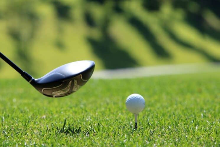 A single golf club hovering over a golf ball perched on a tee on a green lawn