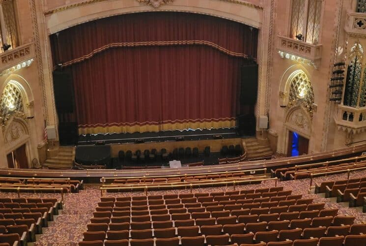 An expansive and ornate theatre looking down from an upper balcony at a stage with red curtains