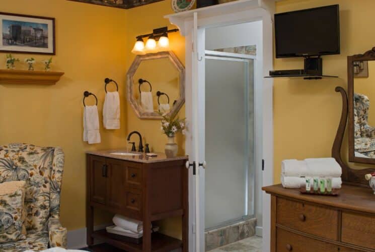 Bedroom with yellow walls, antique dresser with mirror, sink vanity, floral sitting chair, and doorway into bathroom with shower