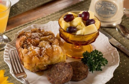 A white breakfast plate with a peach cobbler French toast, sausage patties, cup of fruit, orange juice and mug of coffee