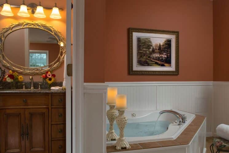 Bathroom with corner jacuzzi tub, two decorative candles, rolled white towels, doorway open showing an elegant vanity with oval mirror