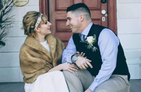 A bride wearing a fur shawl sitting on a porch with her groom in a blue shirt and black vest.