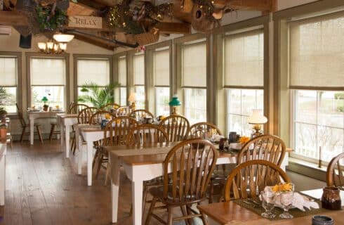 Dining room with several tables with wood chairs, wood beam ceiling, hardwood floors and walls of windows with roman shades