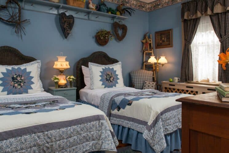 Spacious bedroom with two twin beds, each with colorful blue quilts and pillows, country-chic decor and window with lace curtains