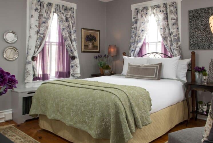 Bedroom with queen bed, sitting chair, side tables with lamp and vase of flowers, three windows with floral and purple sheer curtains