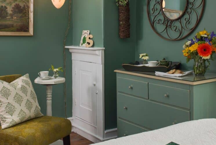 Corner of a bedroom with green walls, sitting chair with pillow, dresser with vase of flowers and tray with teapot and cup