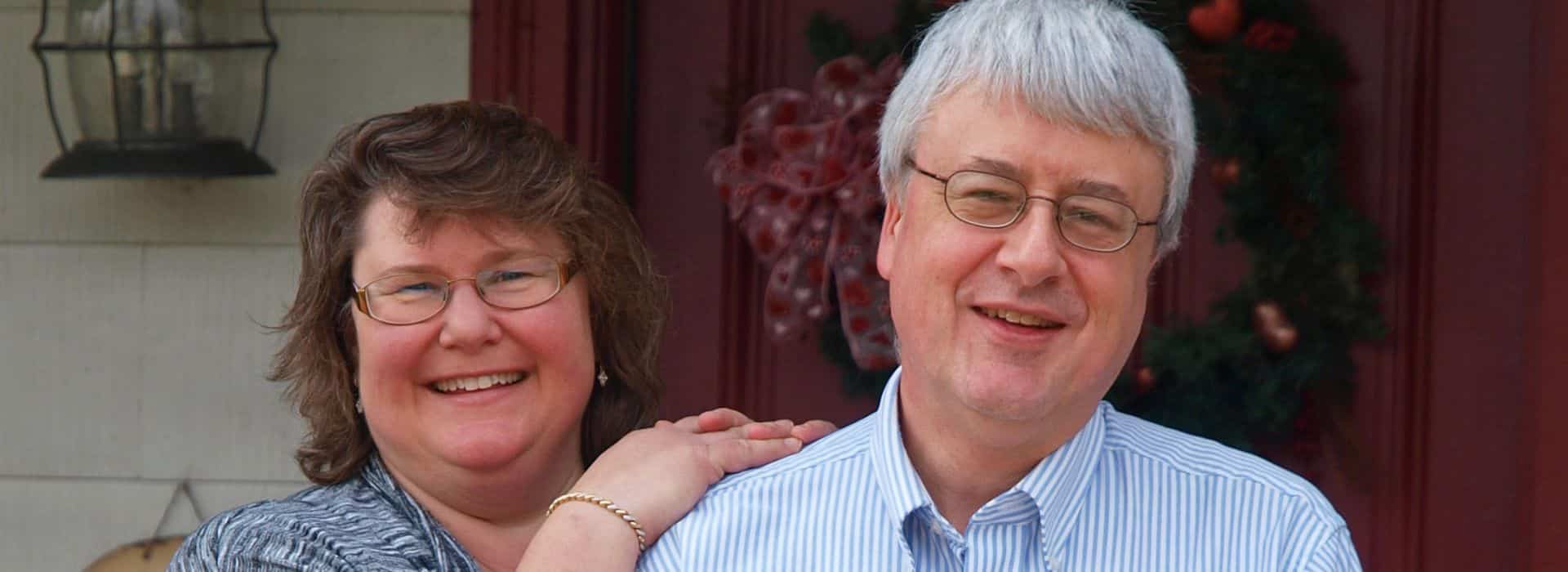 A man with glasses and in a striped shirt standing next to a woman with brown hair in front of the front door of a home.