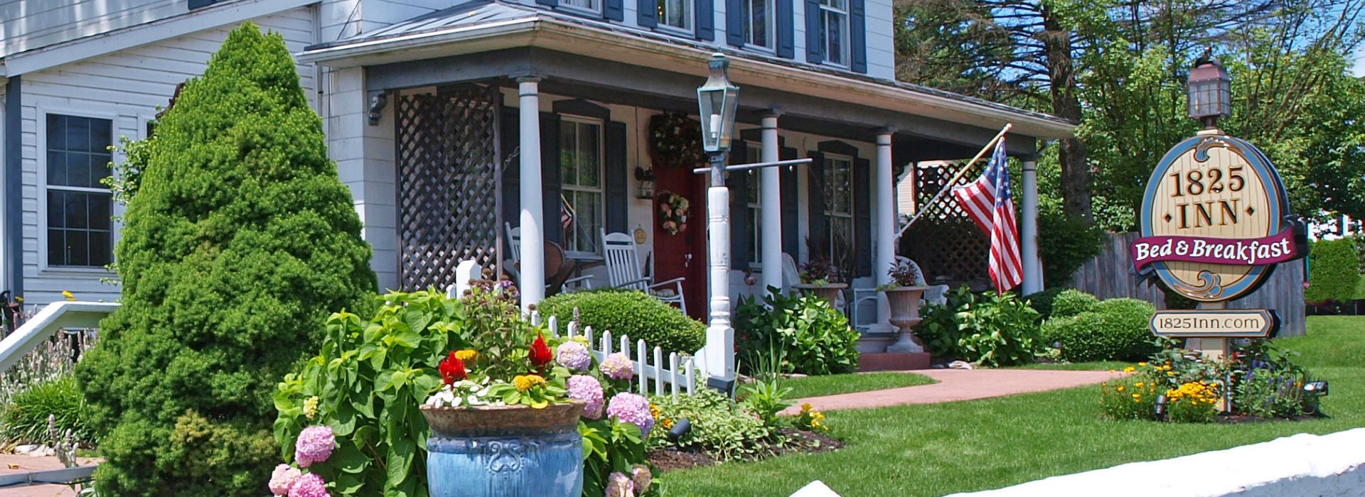 Large oval sign for a bed and breakfast on the front yard of a white home with blue shutters and rocking chairs on the front porch