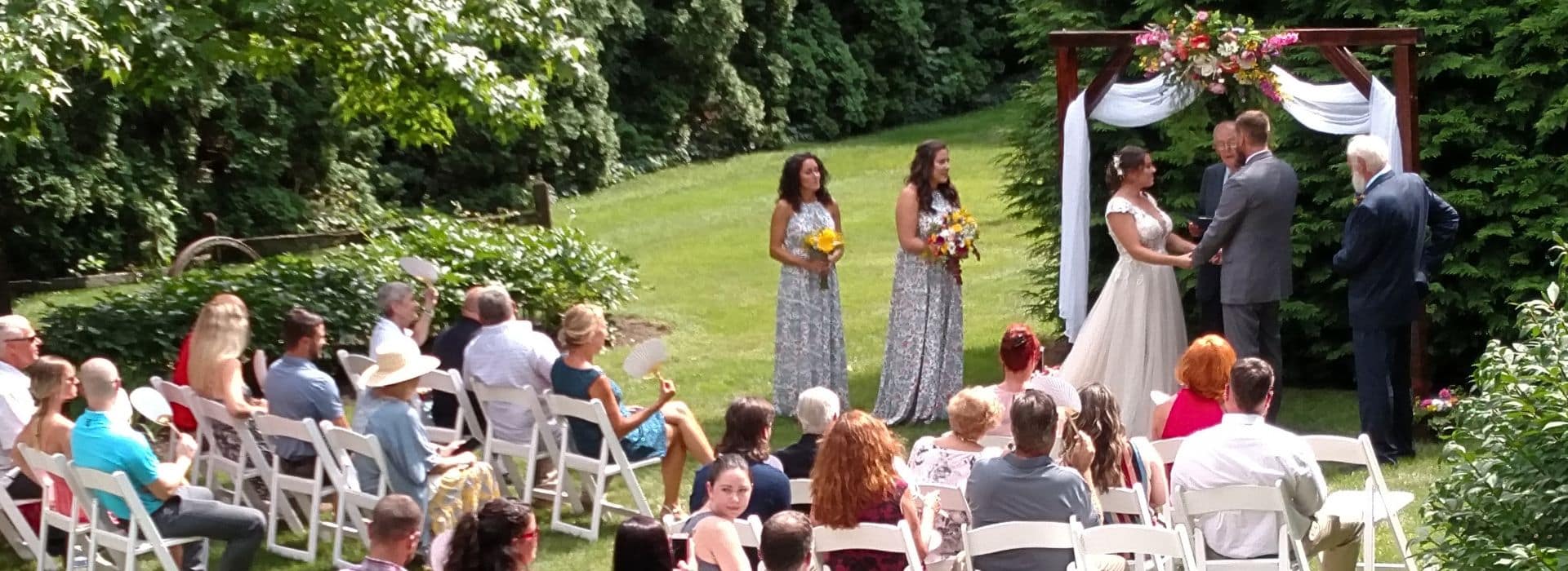 A bride and groom standing outdoors on a lawn surrounded by trees, at a wood arbor with guests in white chairs looking on.