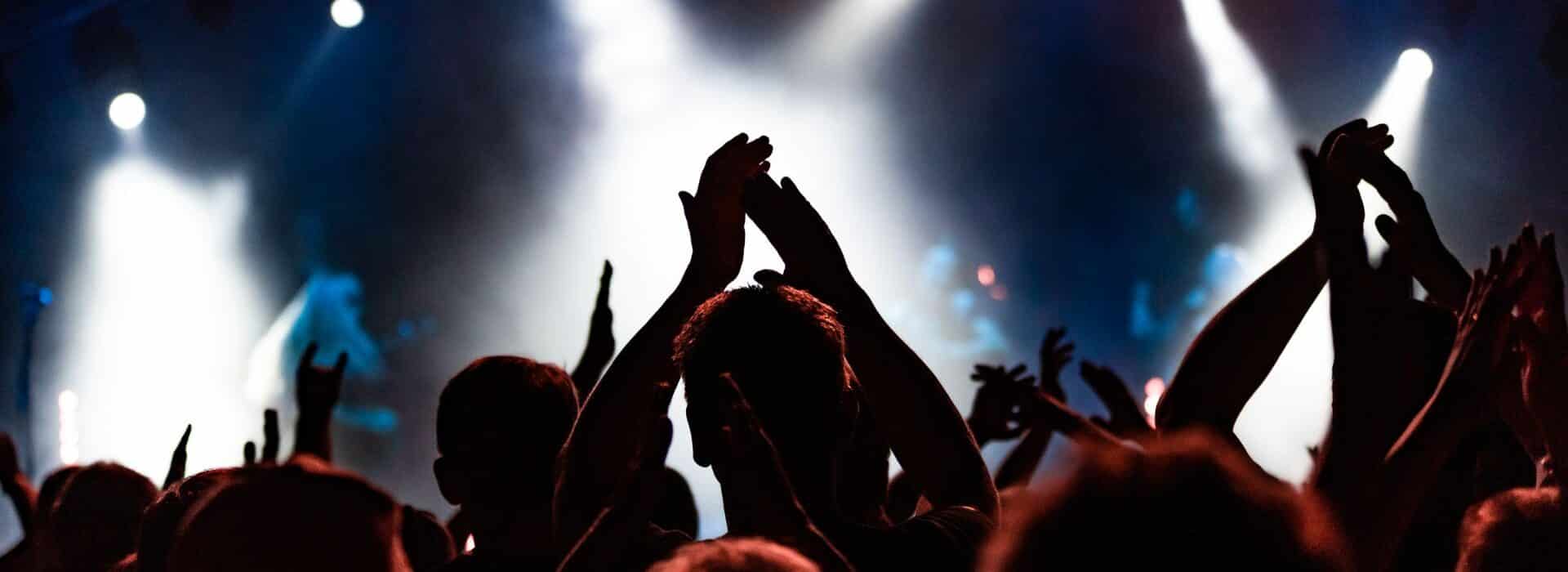 People in a crowd clapping their hands in the air while watching a concert under smoke and lights