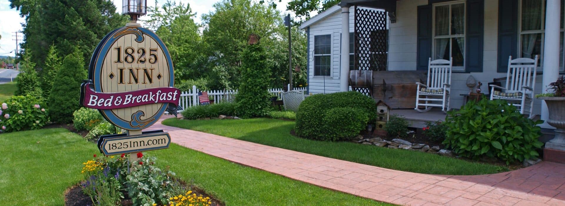 Front facade of a bed and breakfast, porch with two white rocking chairs, lush plants and flowers, sign in front yard