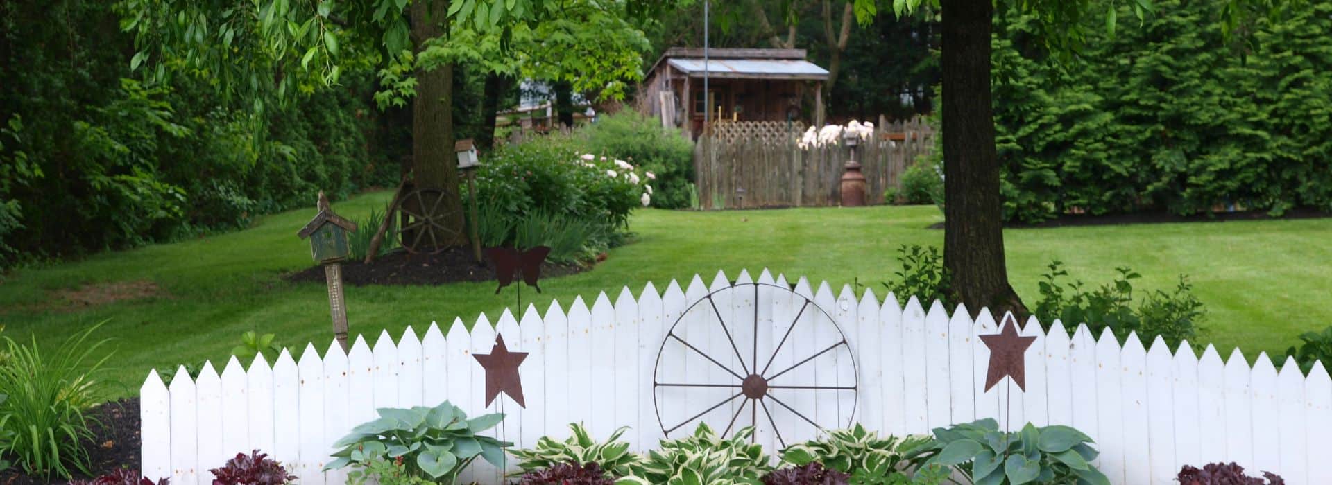 Serene yard with white picket fence in front of lush landscaping and colorful plants, large lawn space lined with trees