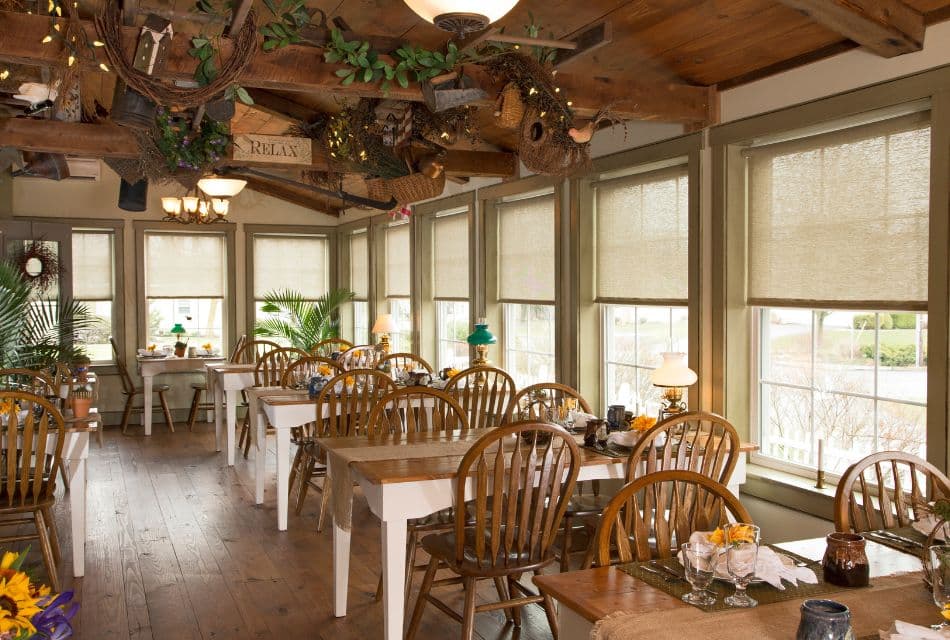 Dining room with several tables with wood chairs, wood beam ceiling, hardwood floors and walls of windows with roman shades