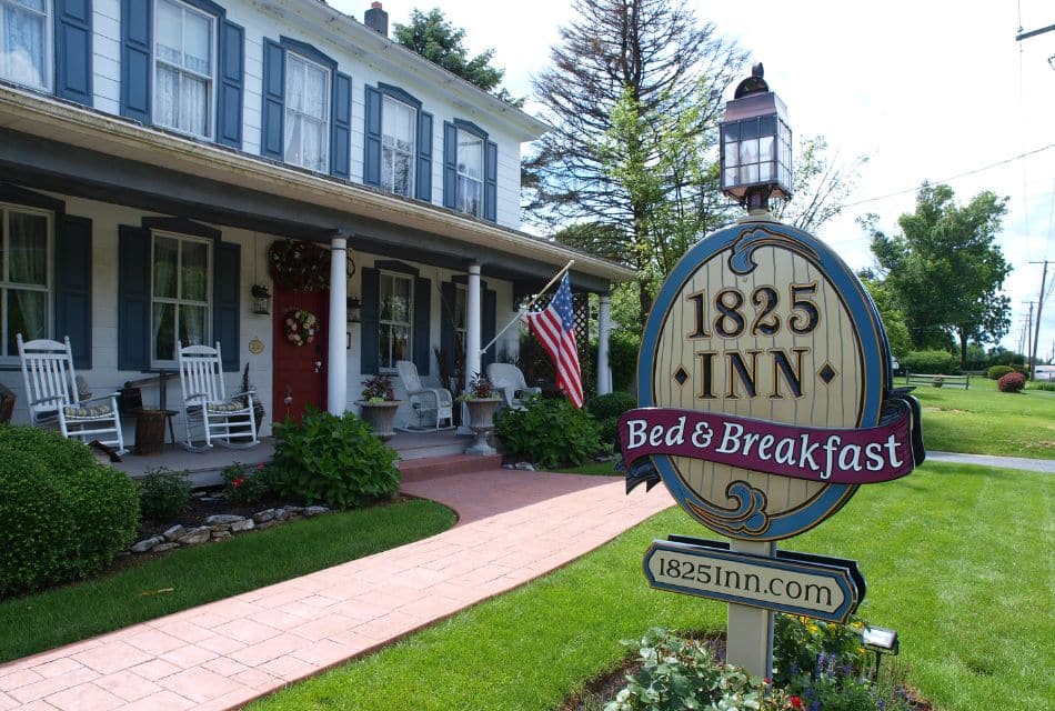 Front facade of an inn with sign on the yard, porch with white rocking chairs and lush landscaping and plants