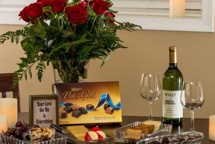 A bouquet of red roses, platters of fruit, cookies and chocolates and a bottle of wine with two glasses on a wood table.